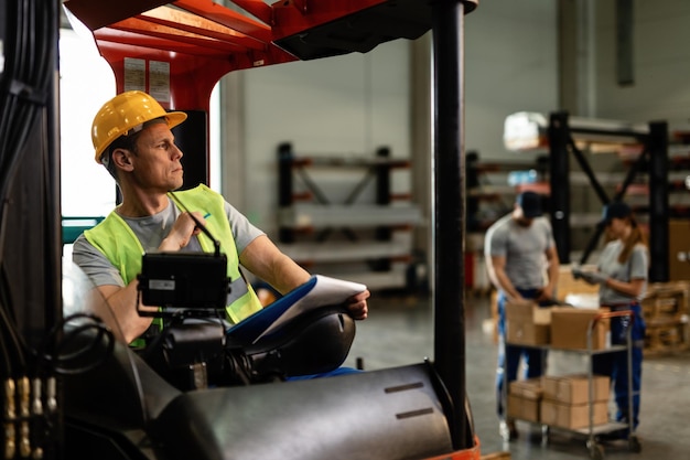 Magazijnheftruckchauffeur die aan iets denkt terwijl hij het leveringsschema in een industrieel gebouw doorloopt