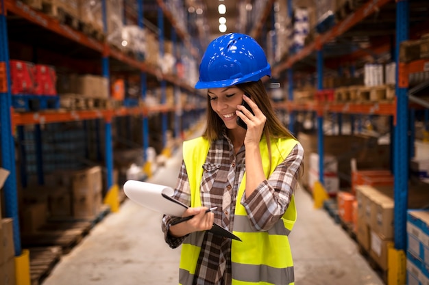Magazijnmedewerker met veiligheidshelm met gesprek aan de telefoon en controlelijst in distributie magazijnfaciliteit
