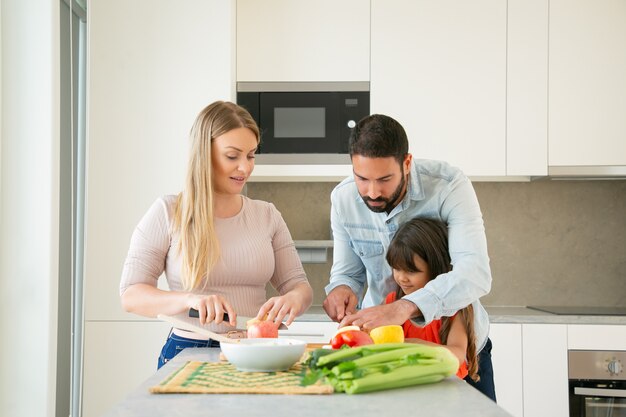 Mama en papa die kind leren koken. Jong koppel en hun meisje vers fruit en groenten voor salade snijden aan de keukentafel. Gezonde voeding of levensstijl concept