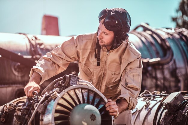 Man bij werkplaats buiten repareert zijn eigen jet. Hij houdt een deel van de turbine vast. De man draagt de helm van de piloot.