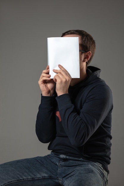 Gratis foto man die gezicht bedekt met boek tijdens het lezen op grijze muur