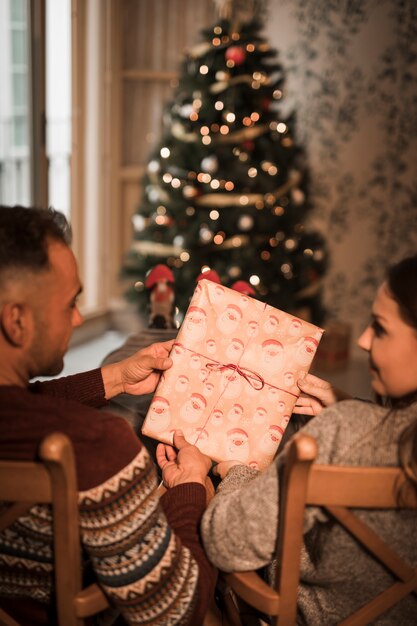 Man en vrouw die huidige doos op stoelen houden dichtbij Kerstboom