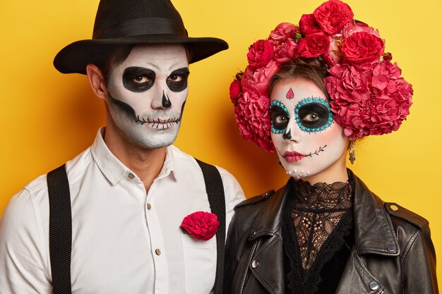 Man en vrouw dragen schedelmake-up, zwart-witte kleding, geïsoleerd op gele achtergrond. Ernstige vampiers vieren samen Halloween