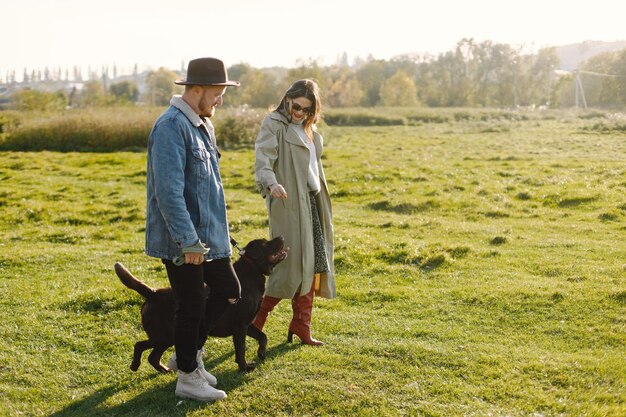 Man en vrouw in modekleren rustend op een natuur met hun labrador. Man met jas en een zwarte hoed en vrouwenrok en rode laarzen