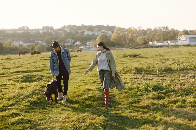 Man en vrouw in modekleren rustend op een natuur met hun labrador. Man met jas en een zwarte hoed en vrouwenrok en rode laarzen