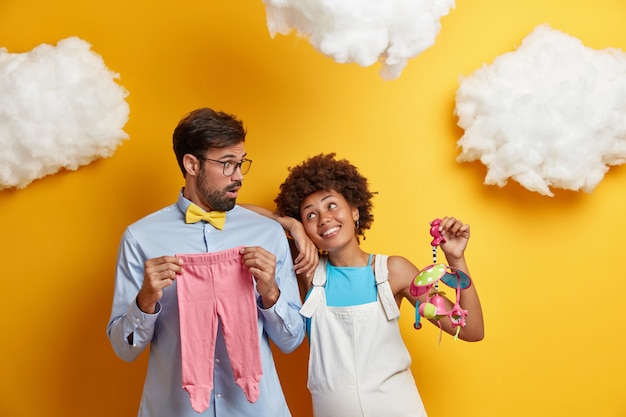 Man en vrouw poseren met babyartikelen en bereiden zich voor om ouders te worden. Vrolijke zwangere vrouw houdt mobiel speelgoed kijkt graag naar man geïsoleerd op gele achtergrond. Ouderschap zwangerschap concept