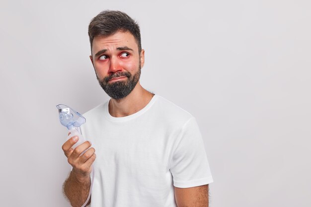 man gebruikt medische apparatuur voor inademing heeft astma-aanval allergische reactie rode gezwollen ogen kijkt verdrietig weg staat op wit