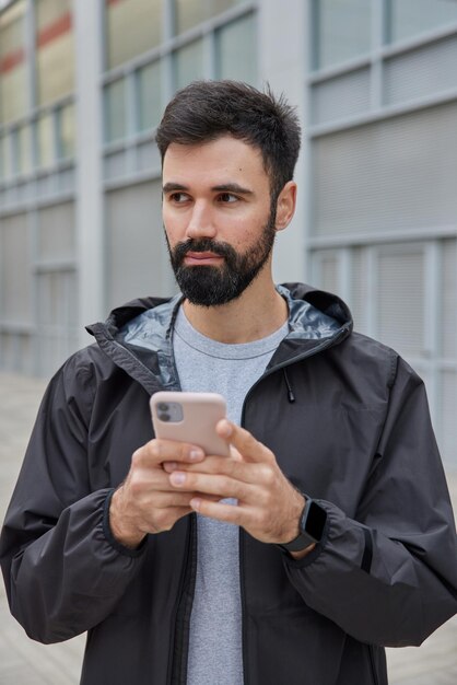 man gekleed in zwarte anorak gebruikt mobiele telefoon om te chatten online downloads applicatie voor sport traning poses buitenshuis op wazig. Moderne levensstijl