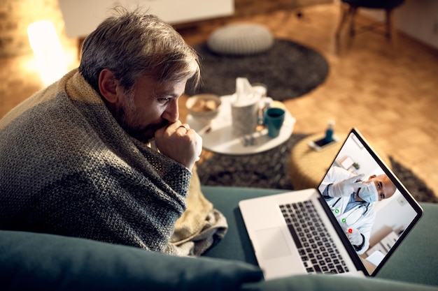 Man gewikkeld in deken die 's avonds thuis videogesprek voert met een dokter