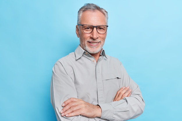 man houdt armen gevouwen kijkt met zelfverzekerde vrolijke uitdrukking draagt formeel shirt en bril voor oogcorrectie op blauwe muur