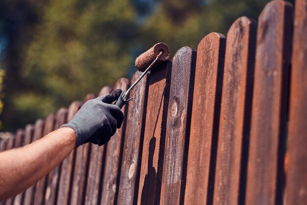 Man in beschermende handschoenen schildert houten hek in heldere zomerdag.