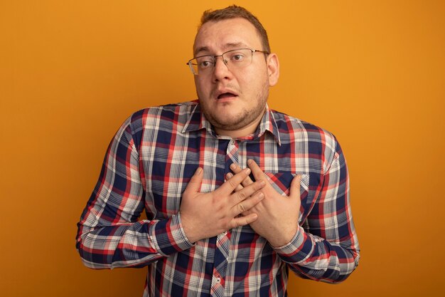 Man in bril en geruit overhemd kijkt opzij met handen op zijn borst bezorgd staande over oranje muur