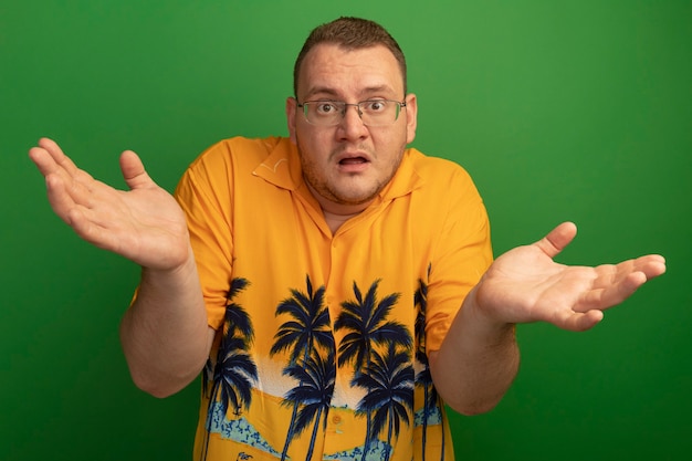 Gratis foto man in bril en oranje overhemd verward met opgeheven handen staande over groene muur