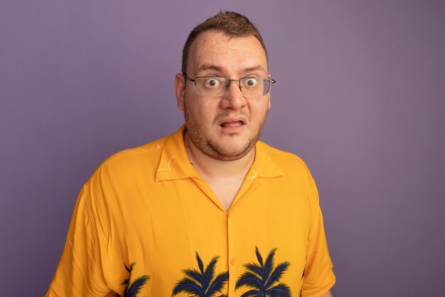 Man in bril met oranje shirt verward en bezorgd staande over paarse muur