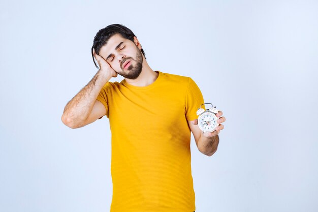 Man in geel shirt met een wekker en ziet er slaperig uit.