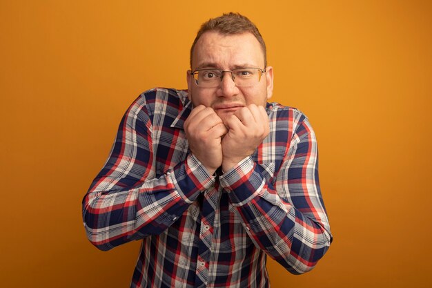 Man in glazen en geruit overhemd beklemtoonde en nerveuze nagels bijten die zich over oranje muur bevinden