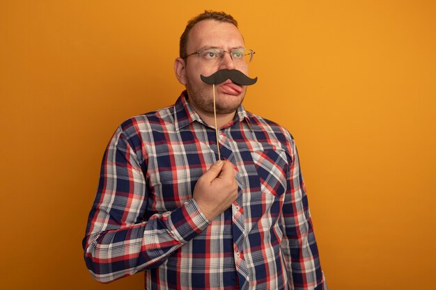 Man in glazen en geruit overhemd die grappige snor op stok houden die uit tong steekt die zich over oranje muur bevindt