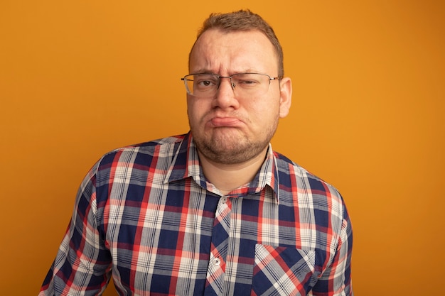 Man in glazen en geruit overhemd met fronsend gezicht dat ontevreden is over oranje muur