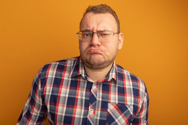 Man in glazen en geruit overhemd met fronsend gezicht dat ontevreden is over oranje muur