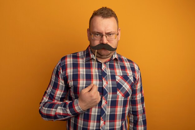 Man in glazen en geruit overhemd met grappige snor op stok die zich over oranje muur bevindt