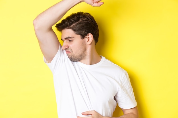 Man in het zweet die zijn oksel ruikt in een wit t-shirt en grimassen trekt van stinkende kleding