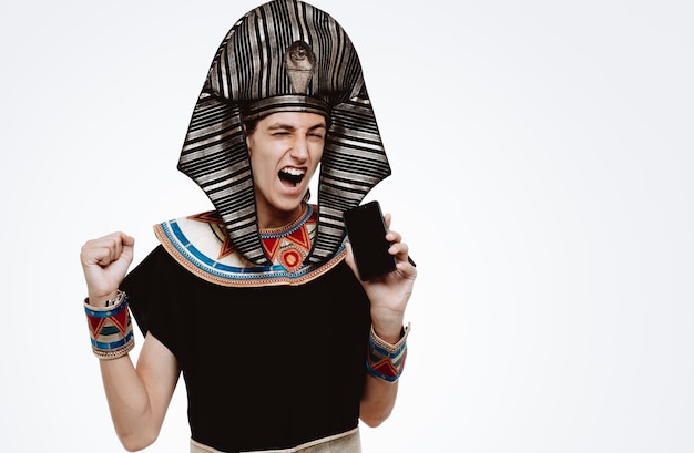 Man in oud egyptisch kostuum met smartphone balde vuist opgewonden en gek blij met zijn succes op wit