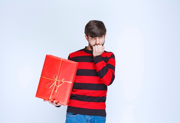 Man in rood gestreept shirt met een rode geschenkdoos en ziet er verward en attent uit.