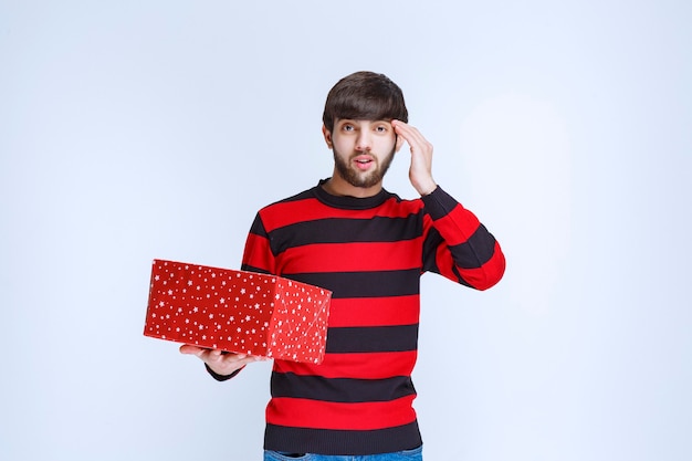 Man in rood gestreept shirt met een rode geschenkdoos ziet er verward en attent uit.