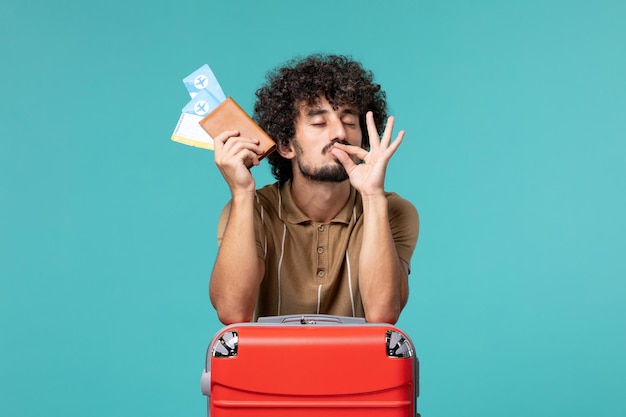 Man in vakantie met kaartjes en leunend tegen zijn rode tas op blauw