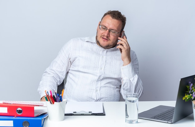 Man in wit overhemd met een bril die op een mobiele telefoon praat met een serieus gezicht aan de tafel met laptop-kantoormappen en klembord op wit