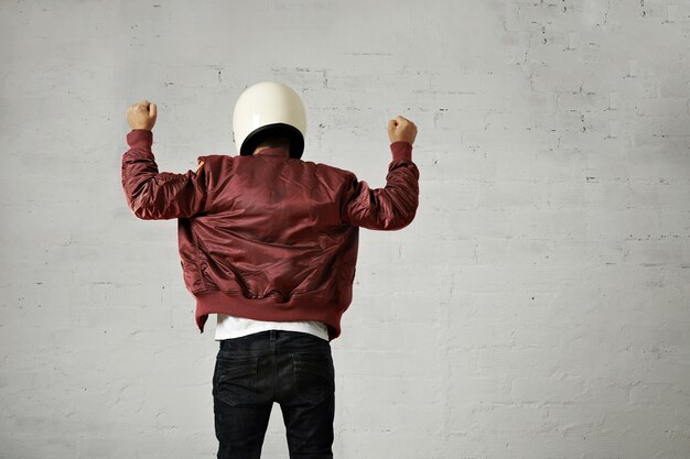 Man in witte motorhelm en bordeauxrode pilotenjas schoot vanaf de achterkant met beide vuisten in de lucht met shaka-gebaar tegen witte muurachtergrond.