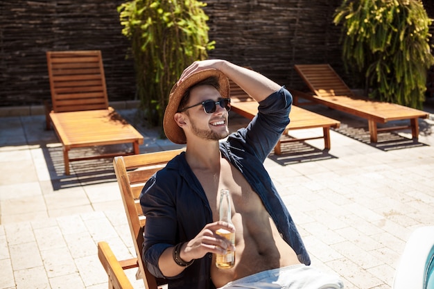 Man in zonnebril en hoed bier drinken, zittend op chaise