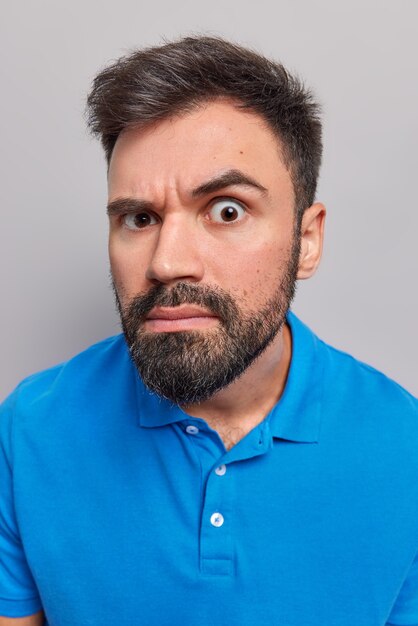 man kijkt aandachtig naar camera heeft nauwgezette uitdrukking draagt casual blauwe t-shirt poses op grijs