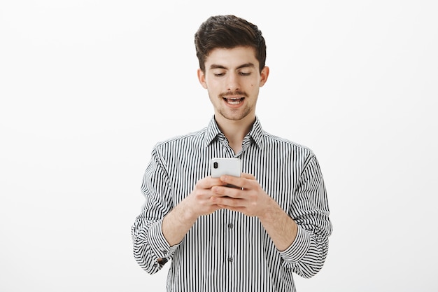 Man leest hardop bericht dat hij op de telefoon heeft ontvangen. Portret van zorgeloze knappe jongeman met baard en snor, smartphone typen, scherm gericht en geïnteresseerd kijken over grijze muur