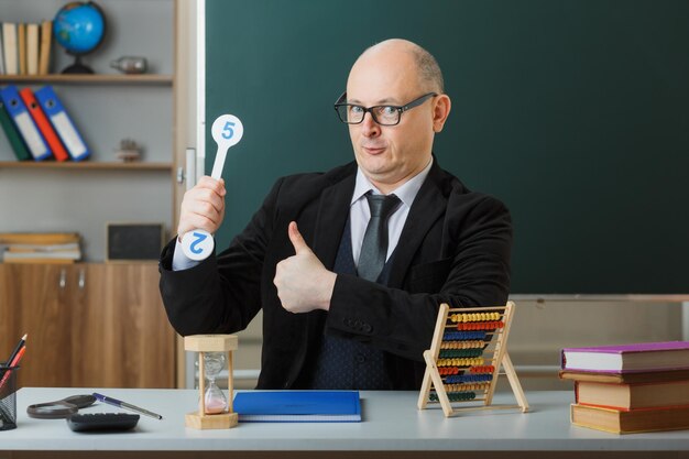 Man leraar met bril zittend aan school bureau met klassenregister voor bord in klas met nummerplaten die les uitleggen die duim omhoog gelukkig en positief toont