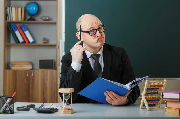 Man leraar met een bril die aan de schoolbank zit met klassenregister voor het bord in de klas en uitleg geeft over de les die er verbijsterd uitziet