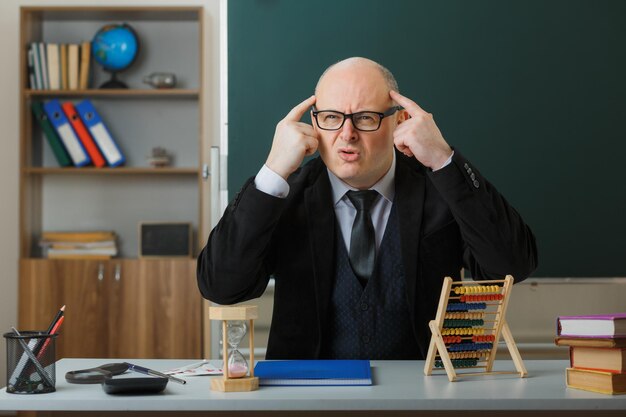 Man leraar met een bril die aan de schoolbank zit met klassenregister voor het bord in de klas en uitlegt dat de les geïrriteerd en geïrriteerd is