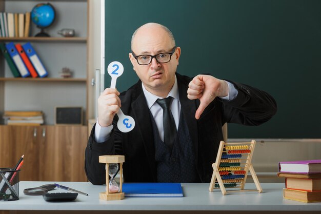 Man leraar met een bril die aan de schoolbank zit met klassenregister voor het bord in de klas met nummerplaten die de les met duim naar beneden uitleggen