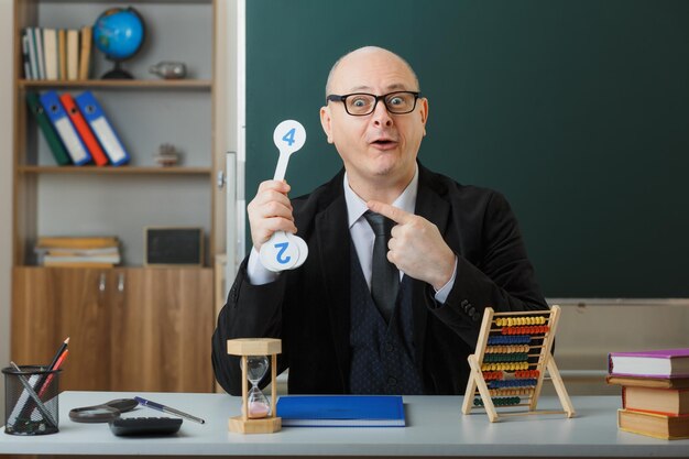 Man leraar met een bril die aan de schoolbank zit met klassenregister voor het bord in de klas met nummerplaten die les uitleggen die wijst met de wijsvinger die er verbaasd uitziet