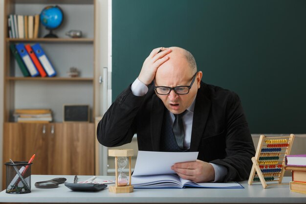 Man leraar met een bril die aan de schoolbank zit voor het bord in de klas en huiswerk controleert van studenten die er geschokt en verward uitzien