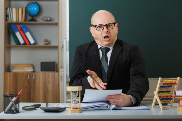 Man leraar met een bril die aan de schoolbank zit voor het schoolbord in de klas en huiswerk controleert van studenten die er ontevreden en verrast uitzien