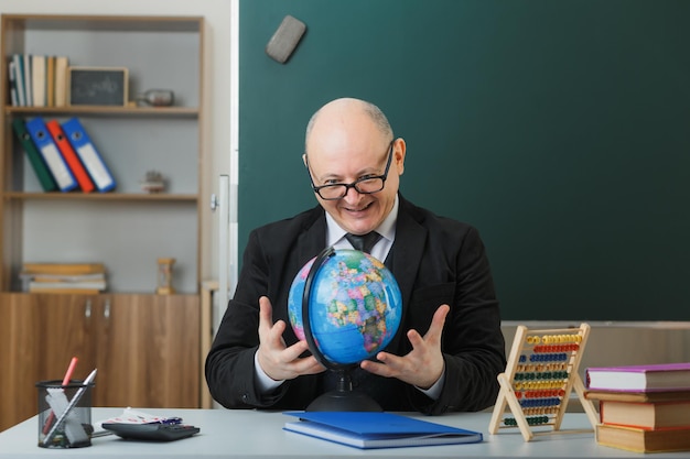 Man leraar met een bril die met een wereldbol op schoolbank zit voor het bord in de klas en de les gelukkig en vreugdevol uitlegt