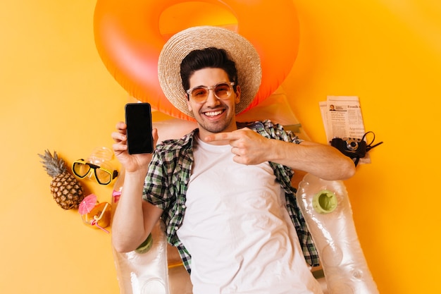 Man ligt op opblaasbare matras op ruimte van ananas, krant en retro camera. man in hoed en bril wijst naar smartphone.