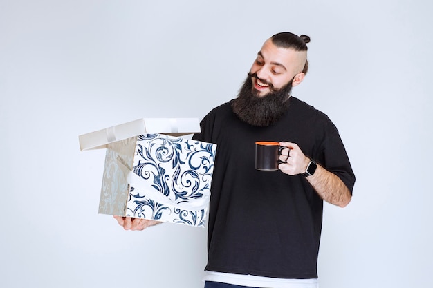 Gratis foto man met baard met een open blauwe geschenkdoos met een kopje koffie en ziet er gelukkig uit.