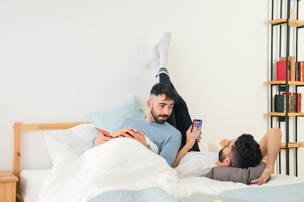 Gratis foto man met boek in de hand te kijken naar zijn vriend met behulp van de mobiele telefoon op bed thuis