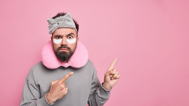 man met dikke baard wijst weg op lege ruimte brengt pleisters onder de ogen aan om rimpels te verminderen draagt een geblinddoekt nekkussen