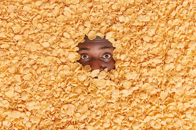 man met donkere huid toont alleen ogen verdronken in granen heeft knapperige snack voor ontbijt maakt creatieve opname van voedselingrediënten. Overhead schot. Cornflakes om te eten