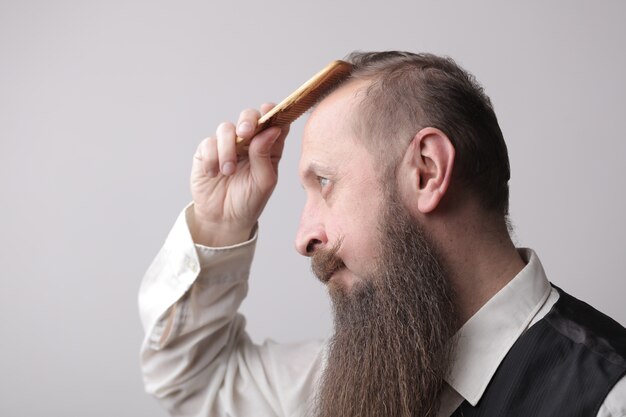 Man met een lange baard en een snor die zijn haar borstelt op een grijze muur