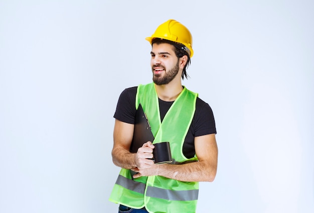 Man met gele helm met projectmap en een kopje koffie.