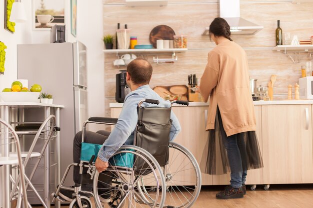 Man met loophandicap in rolstoel die naar vrouw kijkt hoe ze kookt. Gehandicapte verlamde gehandicapte man met loopbeperking integreren na een ongeval.